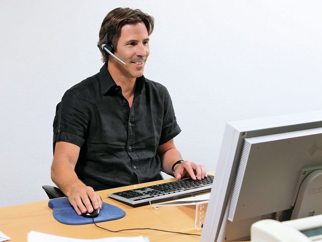 Ein Mann sitzt lächelnd vor dem Bildschirm. Er trägt ein Head-Set, führt ein Telefongespräch und bedient gleichzeitig die Maus.
