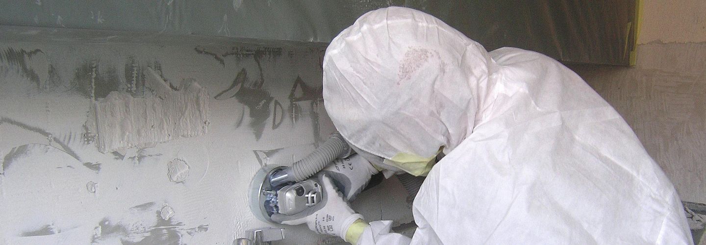 Ein Asbestsanierer entfernt mit einem Schleifgerät den asbesthaltigen Plattenkleber. 