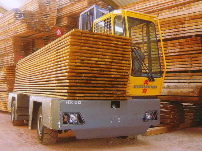 Visione anteriore di un carrello elevatore con una cabina di guida chiusa. Trasporta una grande catasta di pannelli di legno su una piattaforma laterale.