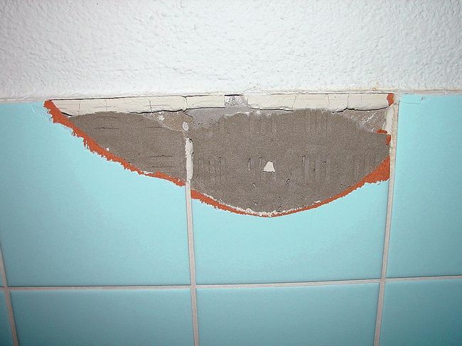 Inquadratura di una parete di un bagno rivestita con piastrelle. È stata staccata una parte di ciascuna delle due piastrelle. Dietro è visibile la colla per piastrelle contenente amianto.