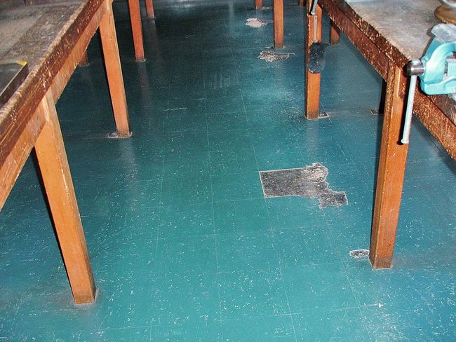 Tra due vecchi banchi da lavoro si vede un rivestimento per pavimenti color petrolio. Manca una piastrella e il rivestimento è rovinato in vari punti.